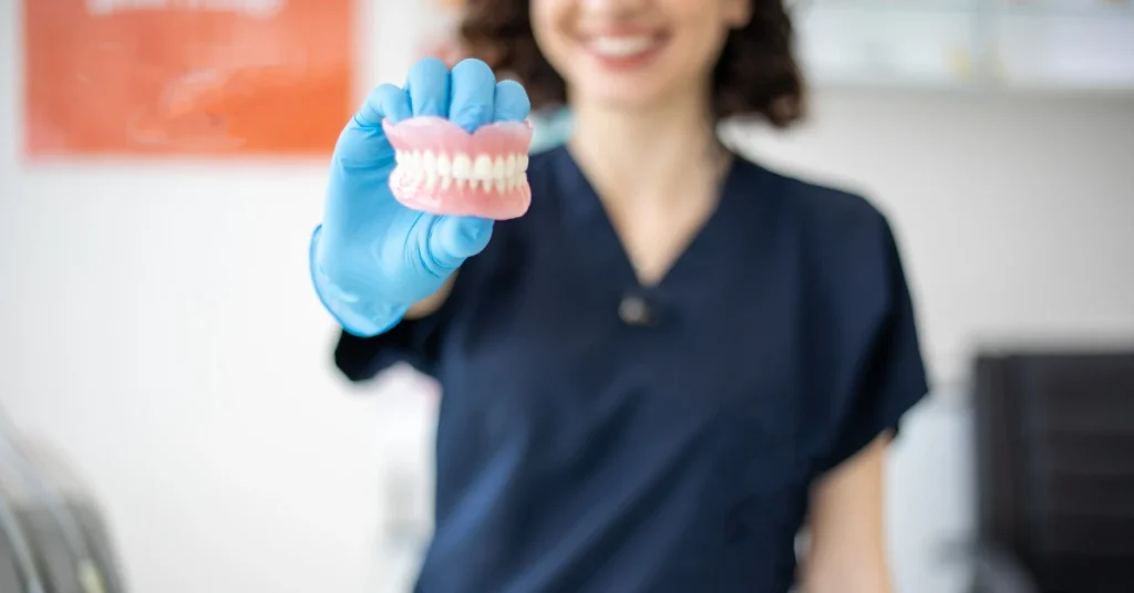 Smiling dental professional in scrubs holding a complete set of dentures with gloved hand in dental office