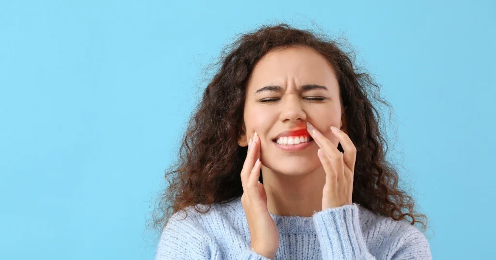 A young woman pressing her cheek with a pained expression, showing discomfort from toothache or gum pain against a blue background.